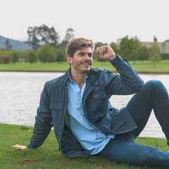 Hombre con chaqueta azul oscuro y camisa azul clara sentado en el césped con lun lago de fondo