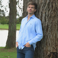 Hombre con camisa azul clara y jeans recostado en un árbol 