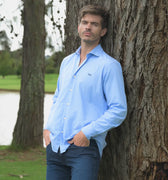 Hombre con camisa azul clara y jeans recostado en un árbol 