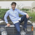 Hombre sonriendo  sentado sobre un carro deportivo con camisa de rayas azul oscuro, jean y botas.