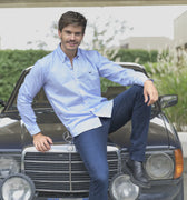 Hombre sonriendo  sentado sobre un carro deportivo con camisa de rayas azul oscuro, jean y botas.