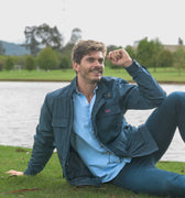 Hombre sentado en un jardin sonriendo con chaqueta azul oscuro, camisa y jeans azules. 